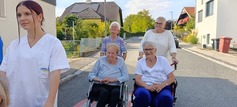 Zwei Bewohnerinnen im Rollstuhl und zwei ehrenamtlich Tätige beim Spaziergang. Zwei ehrenamtlich Tätige schieben Rollstuhl. Praktikantin schiebt Rollstuhl.