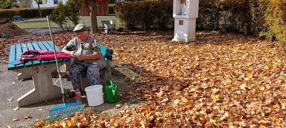 Bewohnerin sitzt auf Bank im Garten. Viel Laub auf Boden.