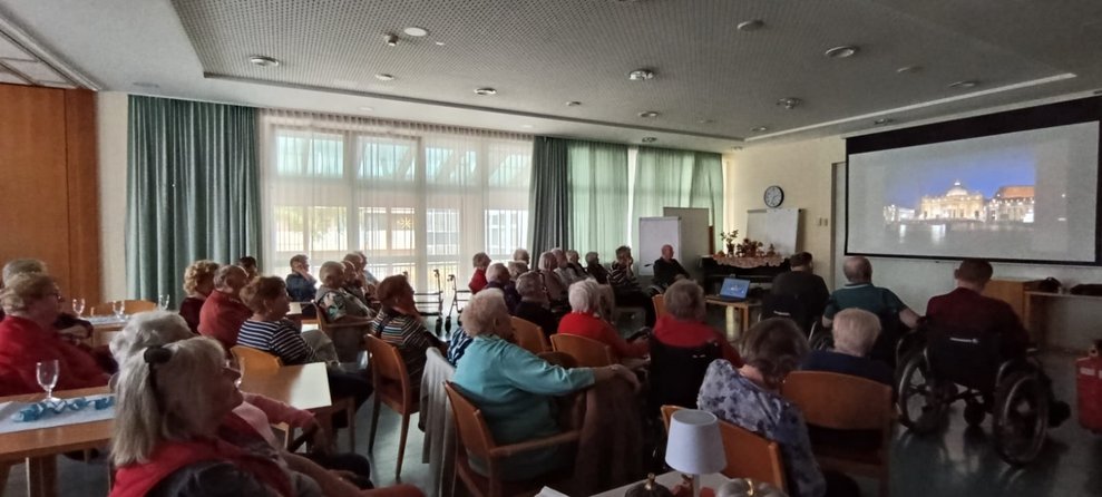 Viele sitzende BewohnerInnen schauen auf Leinwand mit Foto.