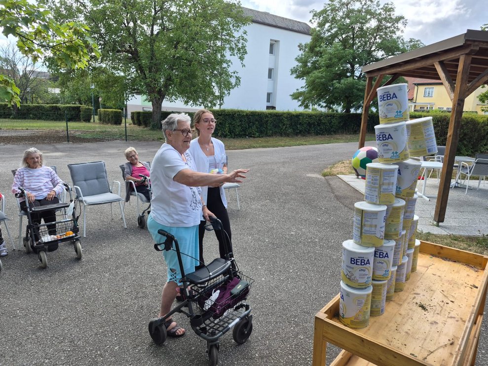 Bewohnerin beim Dosenschießen. Ball berührt Dosen. Dosen wackeln. Praktikantin steht daneben und hält Ball in der Hand.