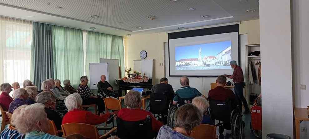Viele sitzende BewohnerInnen schauen auf Leinwand mit Foto. Ehrenamtlich Tätiger steht bei Leinwand.