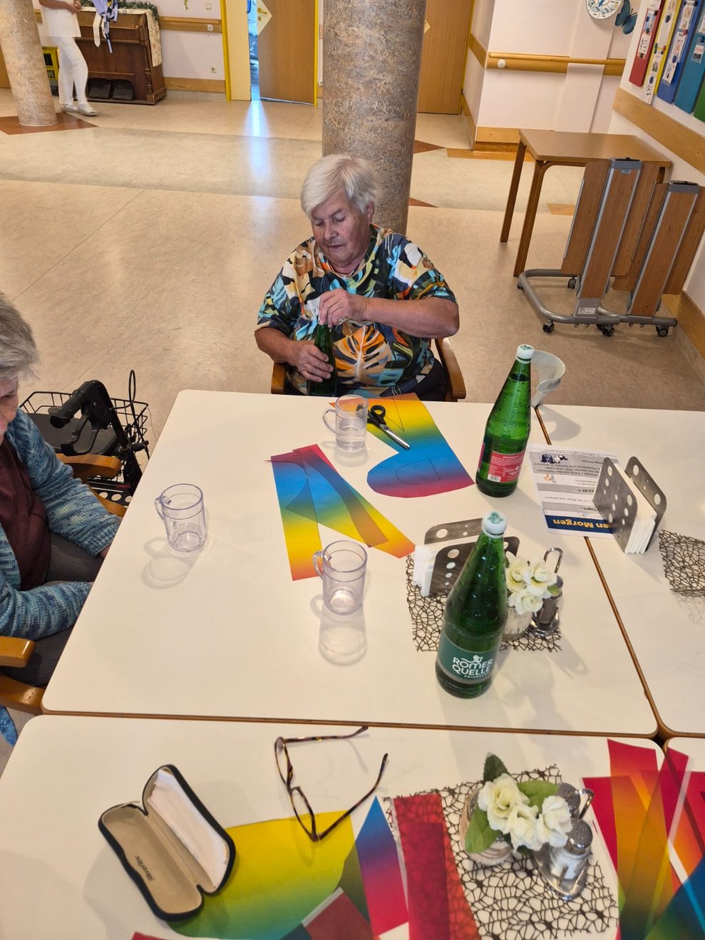 Buntpapier liegt auf Tisch. Bewohnerin öffnet Wasserflasche.
