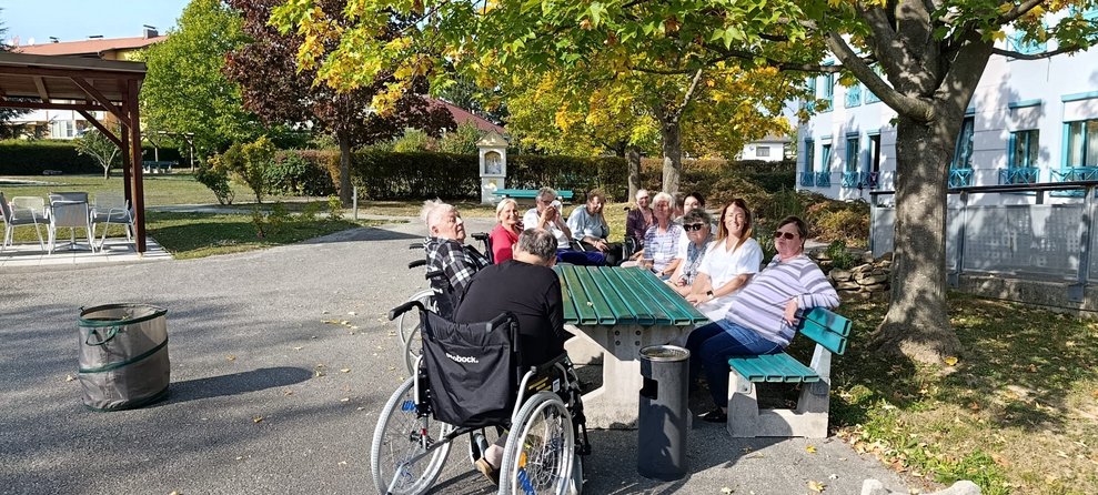Personen sitzen im unserem Garten auf Bank. Tisch in der Mitte. BewohnerInnen im Rollstuhl.