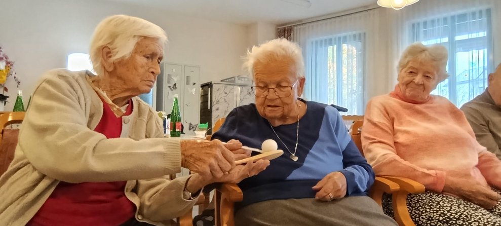 Zwei sitzende Bewohnerinnen halten Kochlöffel in der Hand und kleiner Ball liegt auf Kochlöffel. Eine Bewohnerin sitzt daneben und schaut zu.
