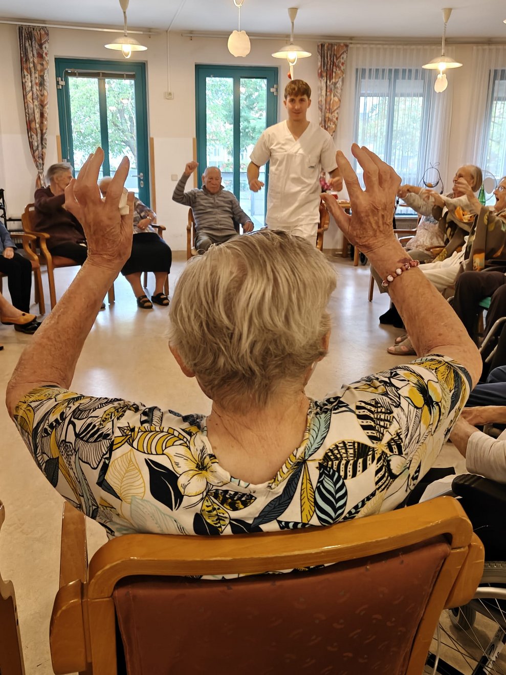 Ferialpraktikant steht in der Mitte zeigt Turnübung vor. Sitzende BewohnerInnen strecken die Hände in die Höhe.