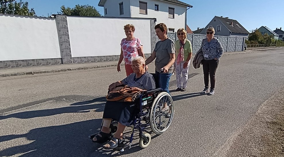 Personen gehen spazieren. Ehrenamtlich Tätige schiebt Rollstuhl mit Bewohner.