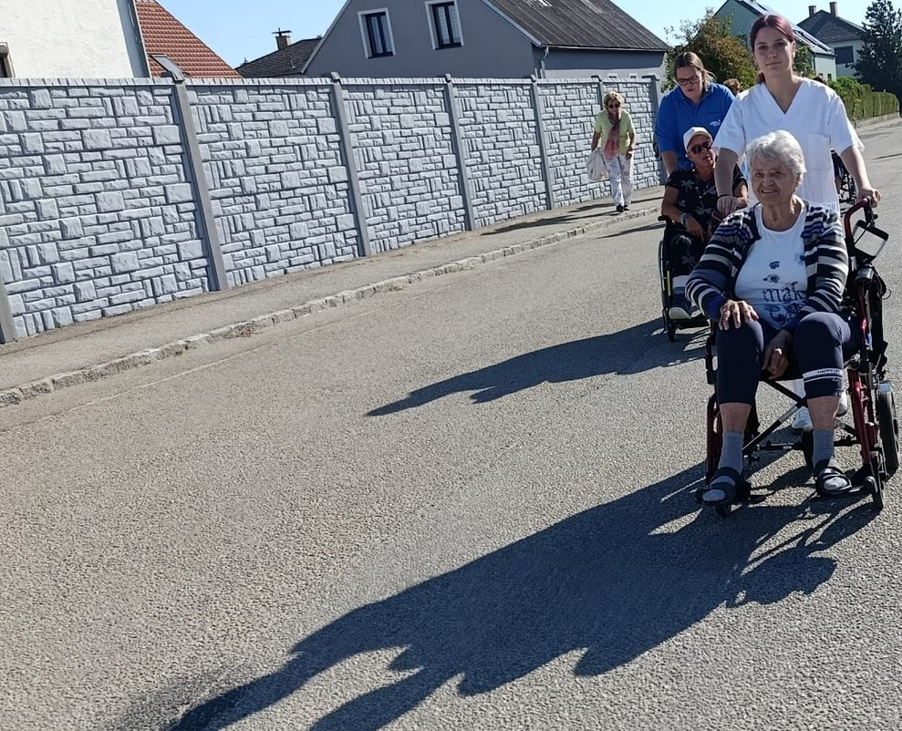 Person geht spazieren. Praktikantinnen schieben Rollstuhl mit Bewohnerinnen.