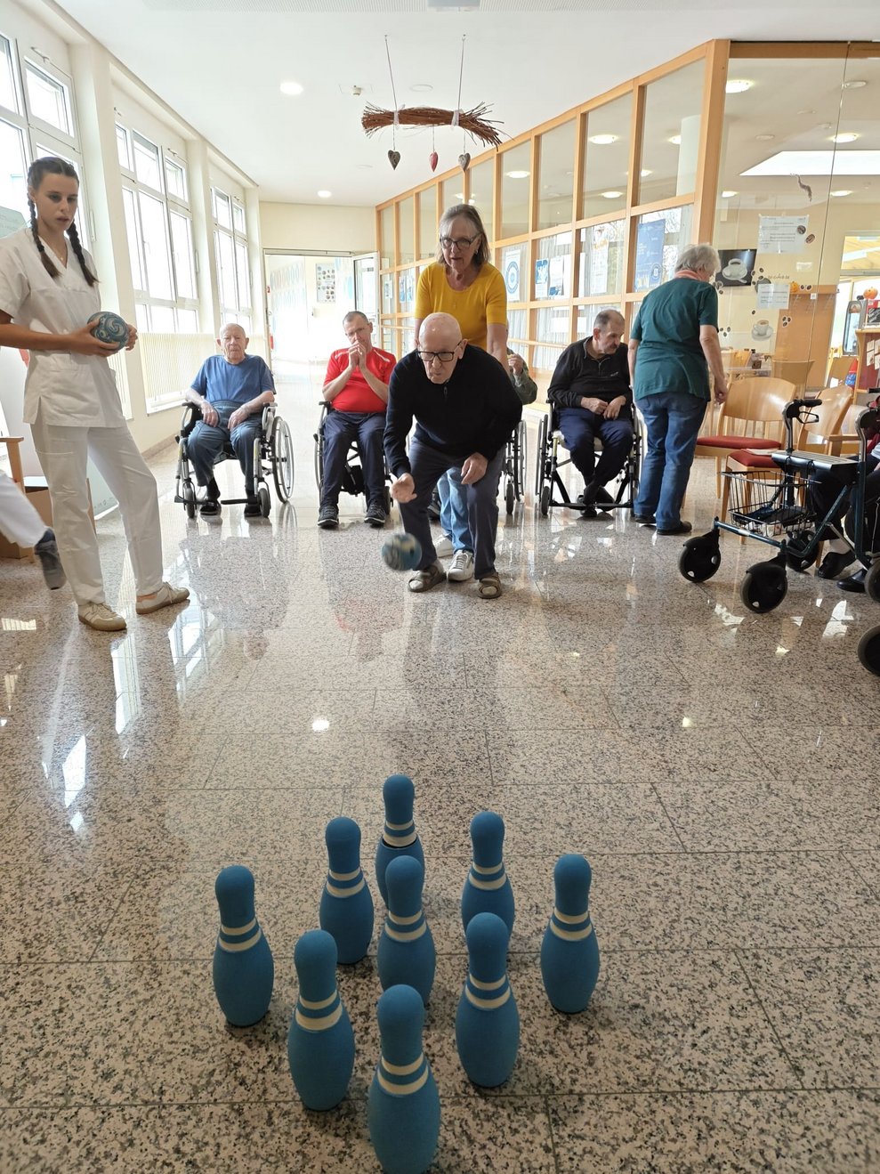 Ehrenamtlich Tätige hält stehende Bewohner beim Kegeln. Ball ist in der Luft und Kegeln auf Boden.