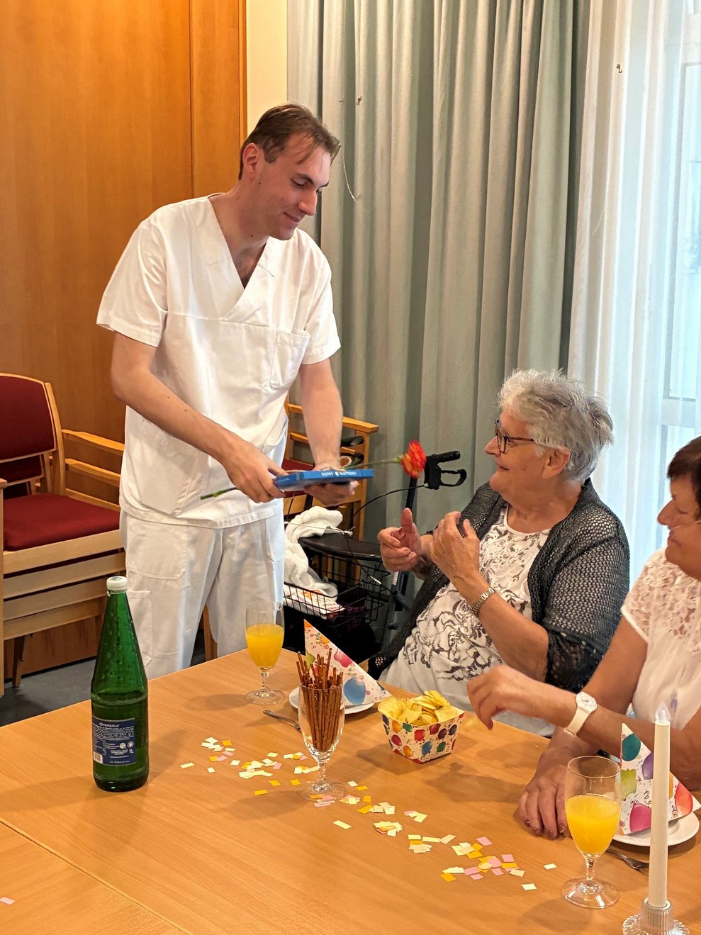 Alltagsbegleiter hält Milka Pralinen Packung und eine Nelke in der Hand und gratuliert Bewohnerin zum Geburtstag.