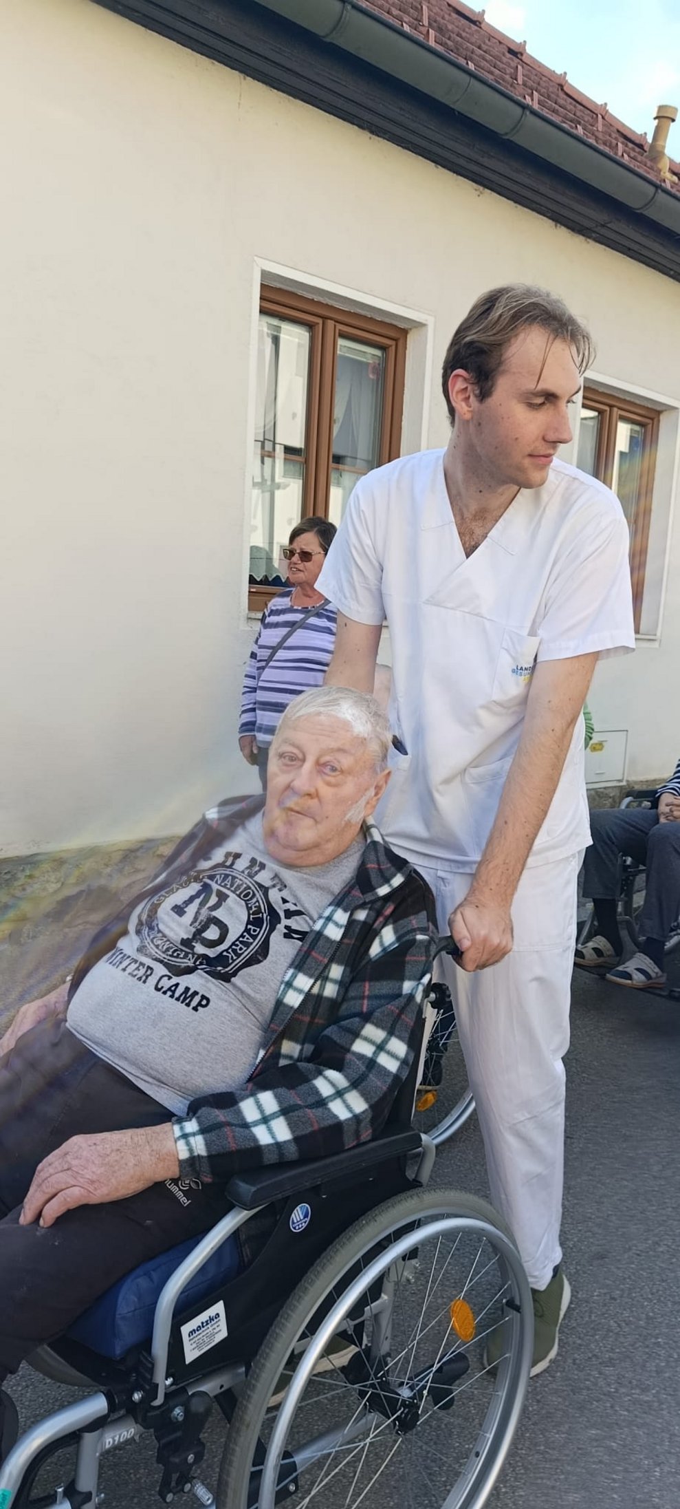 Mann (Alltagsbegleiter) schiebt Rollstuhl mit Bewohner beim Spaziergang.
