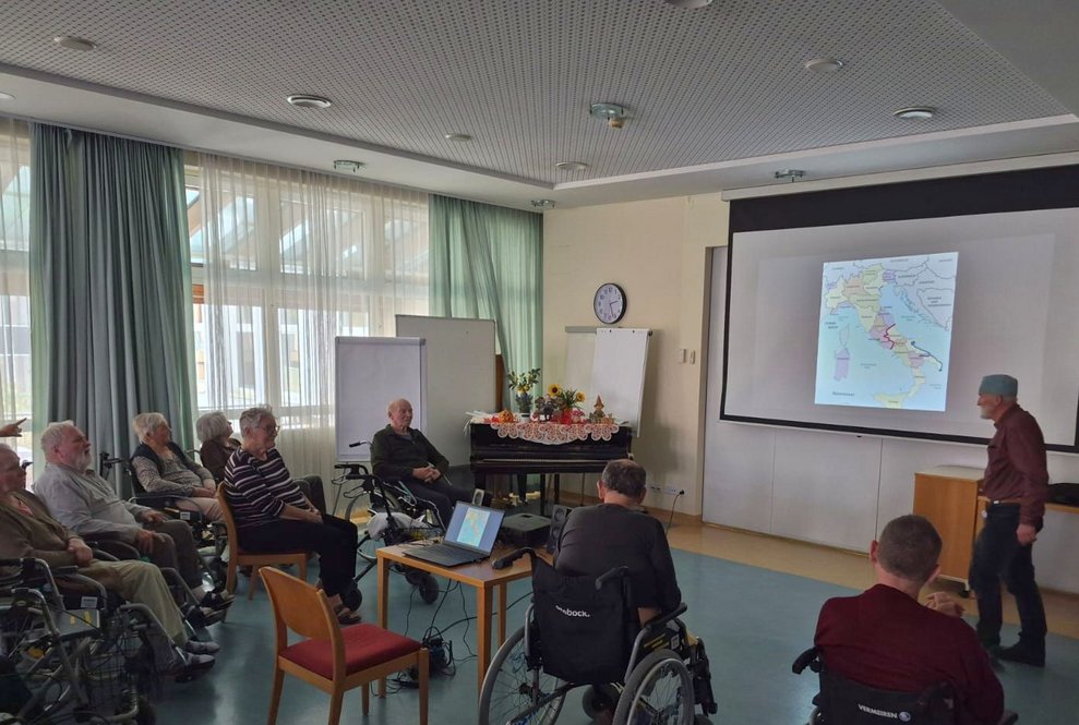 Viele sitzende BewohnerInnen schauen auf Leinwand mit Foto. Ehrenamtlich Tätiger steht bei Leinwand.