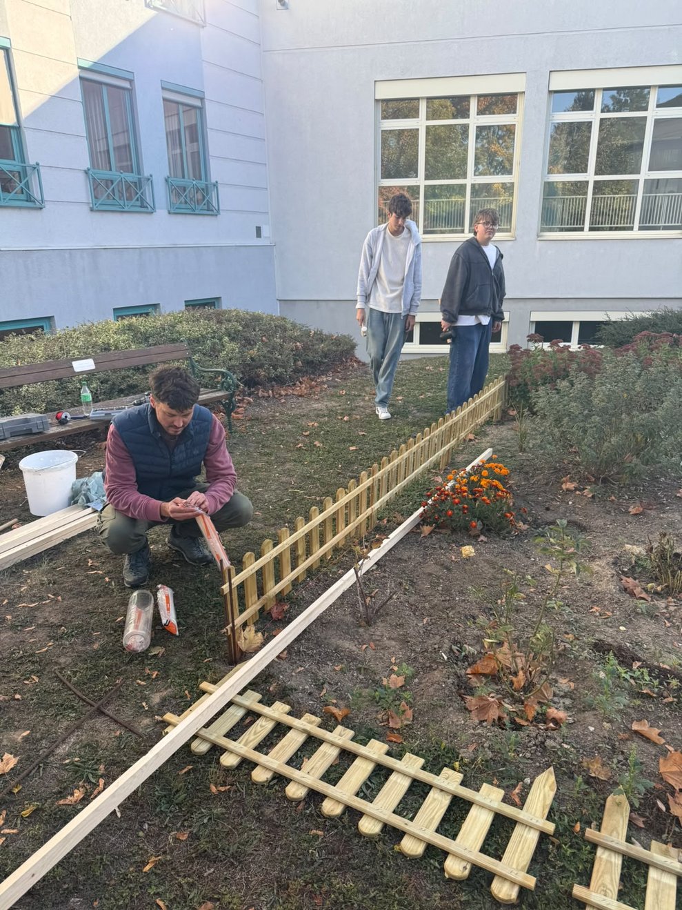 Lehrer und zwei Schüler stellen Holzzaun um das Blumenbeet in unserem Garten auf.