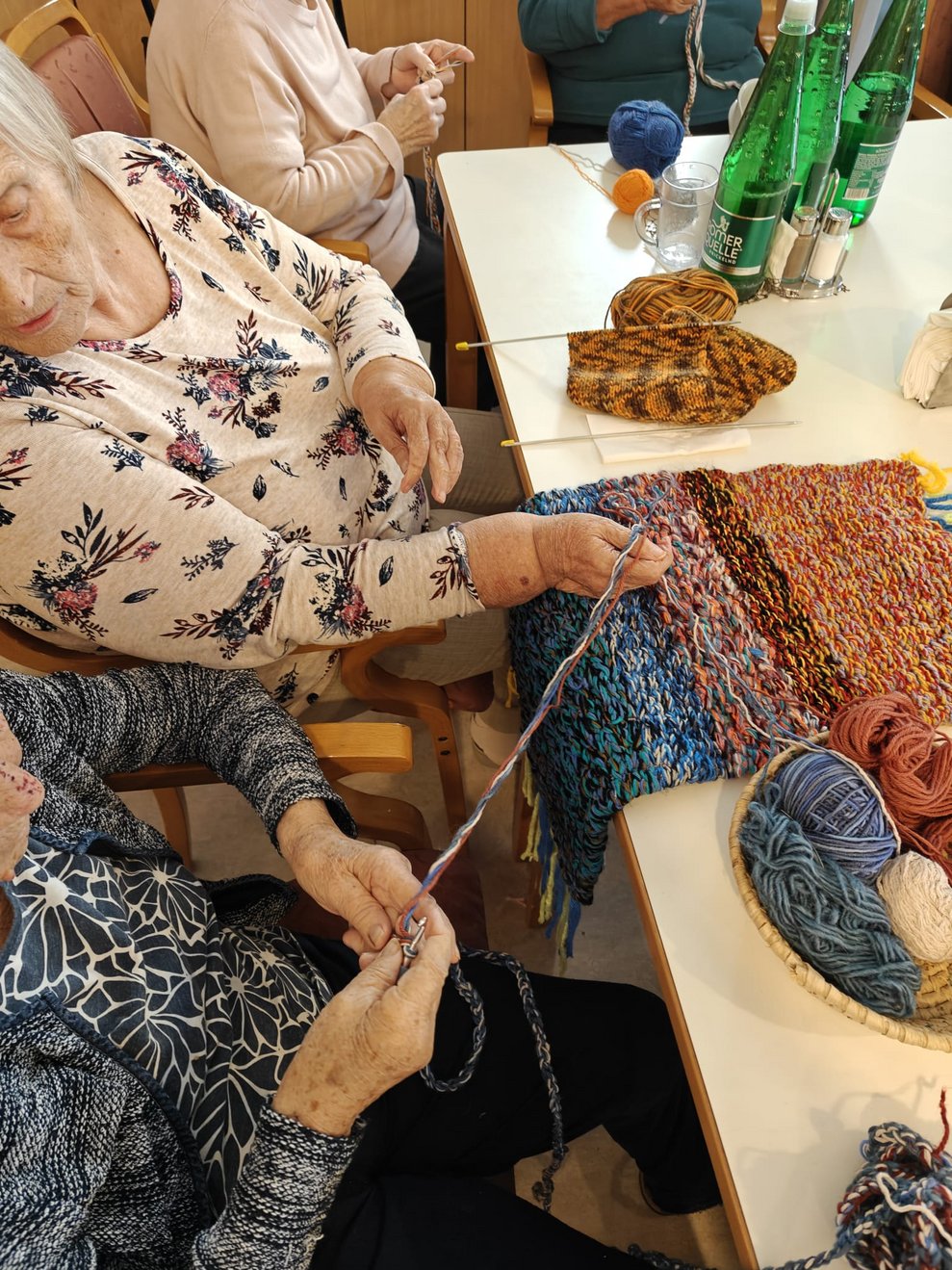 Hände beim Häkeln. Bewohnerinnen halten Wollfaden. Wollteppich liegt auf Tisch.