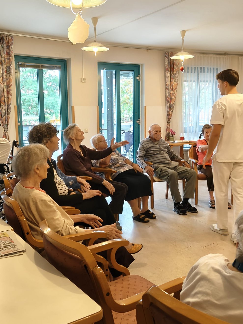 Ferialpraktikant steht. Sitzende BewohnerInnen. Bewohnerin streckt Hand in die Höhe.