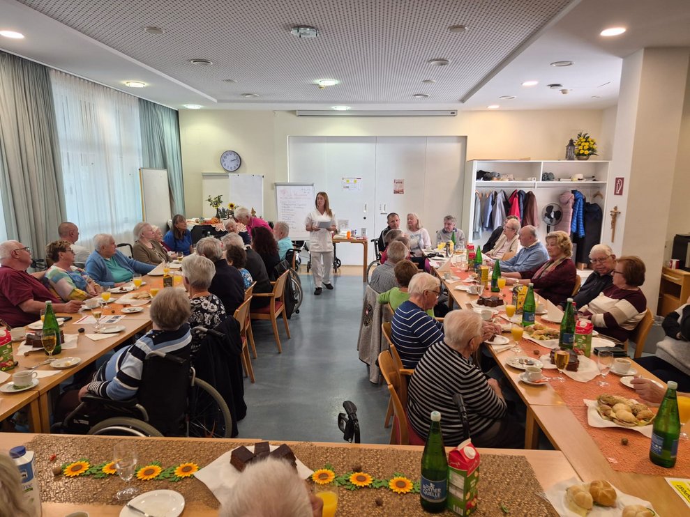 Frau steht und redet. Sitzende Personen beim Tisch.