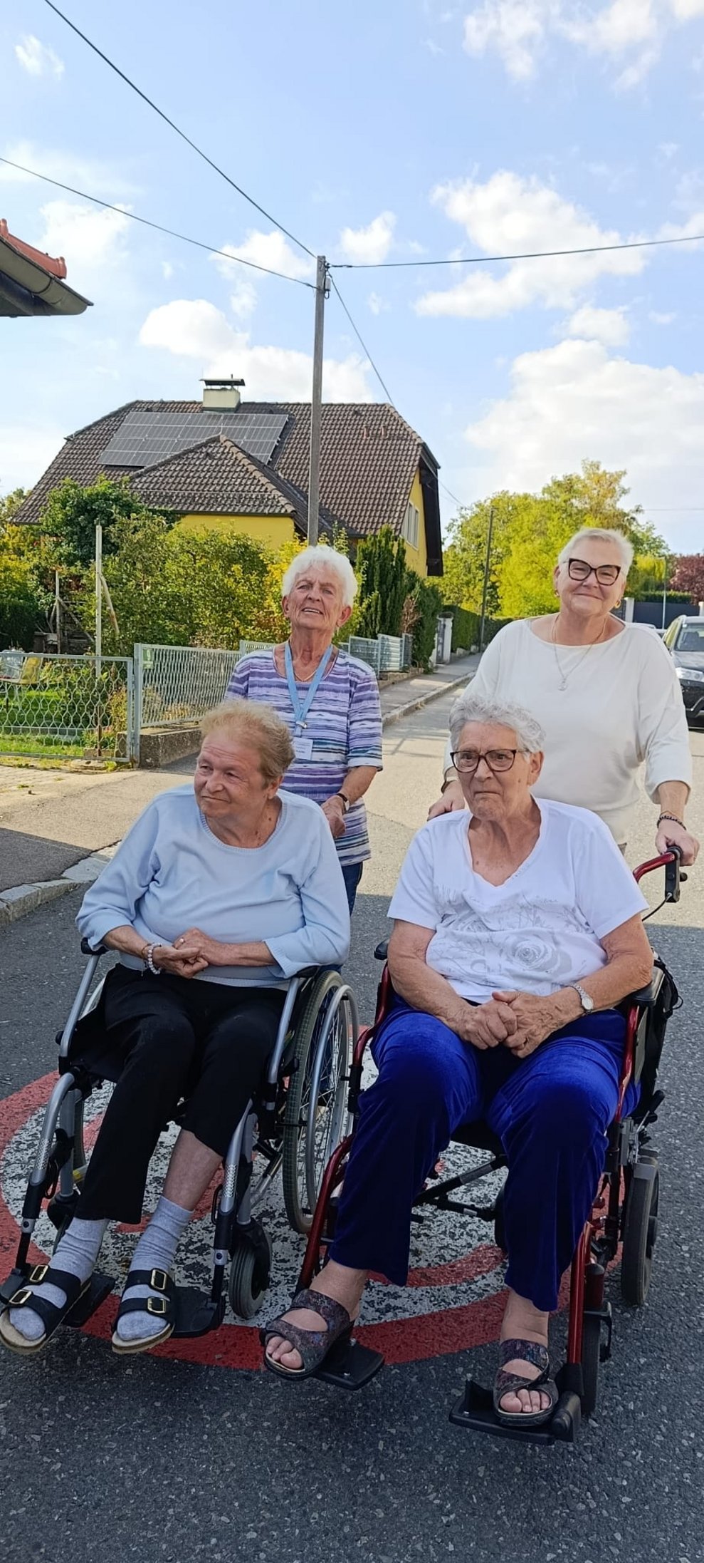Zwei Bewohnerinnen im Rollstuhl und zwei ehrenamtlich Tätige beim Spaziergang. Zwei ehrenamtlich Tätige halten Rollstuhl.