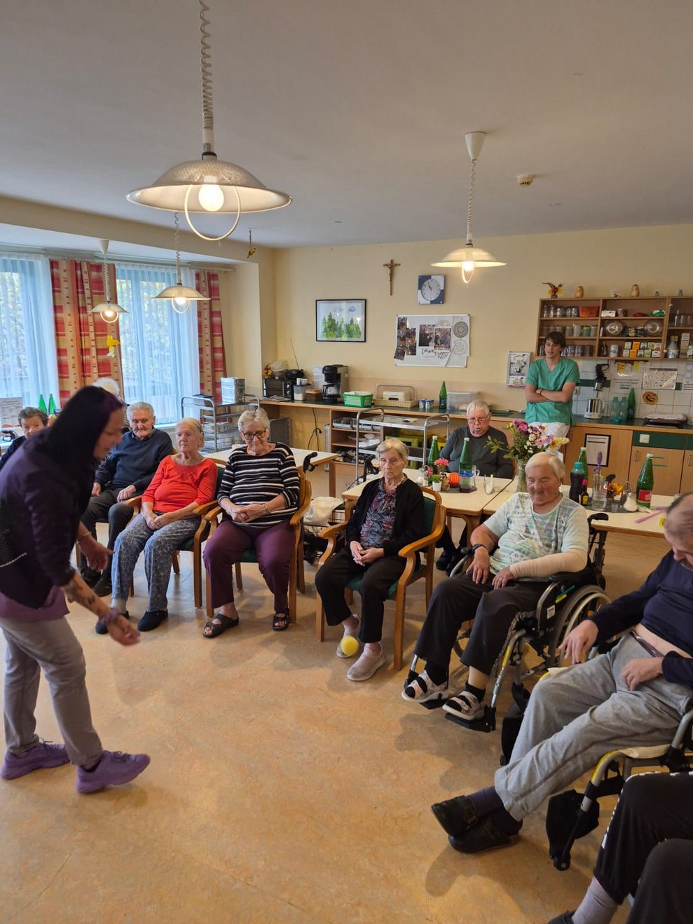 Sitzende BewohnerInnen bei der Bewegungsrunde. Alltagsbegleiterin steht gebückt und versucht kleinen Ball zu fangen.