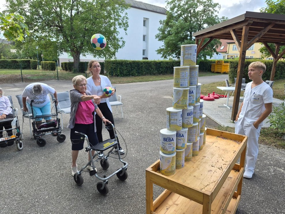 Bewohnerin beim Dosenschießen. Ball in der Luft. Praktikantin steht daneben und hält Ball in der Hand. Mann (FSJ) steht.