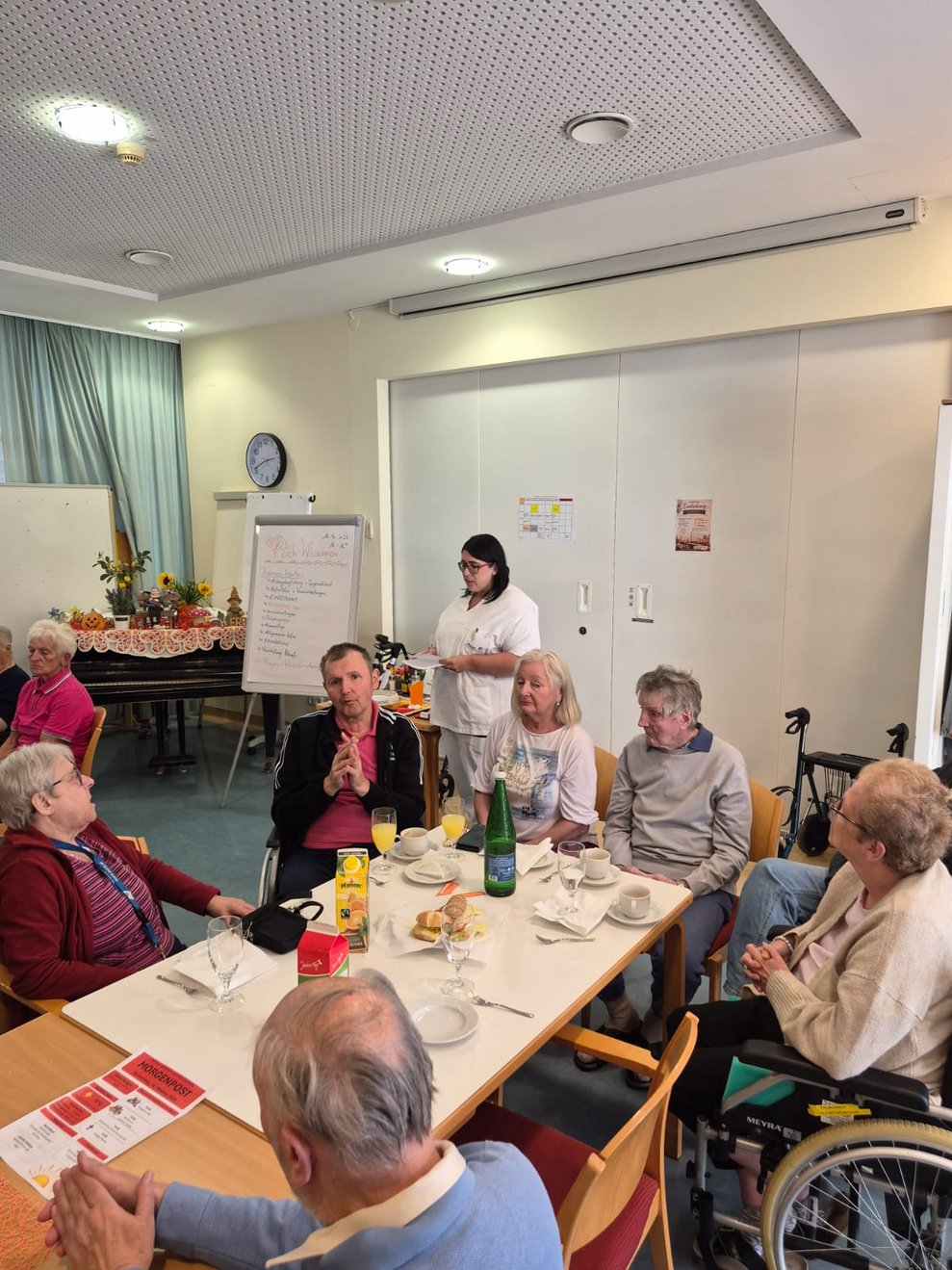 Frau steht und redet. Sitzende Personen beim Tisch.