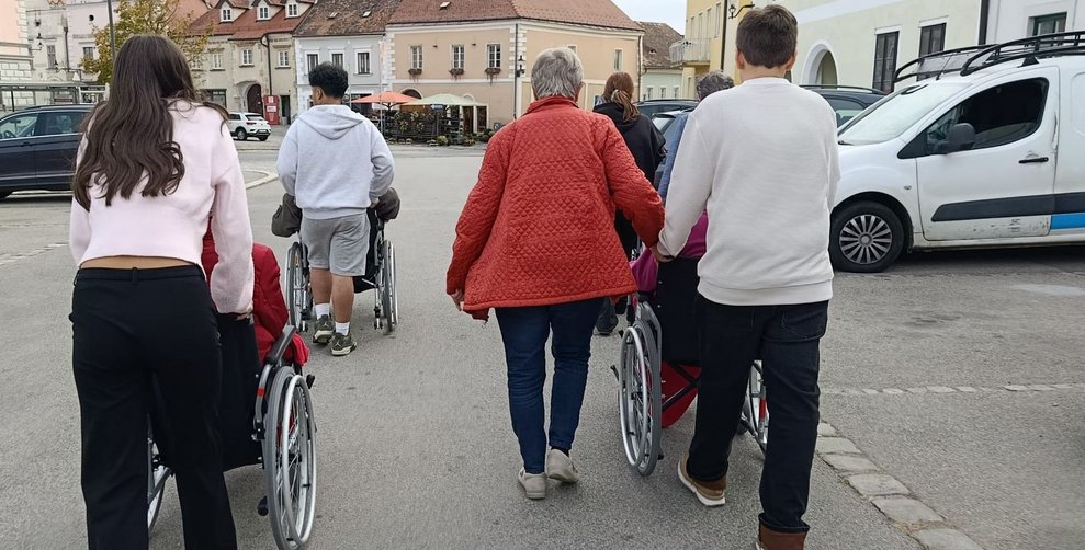 SchülerInnen schieben Rollstuhl mit BewohnerInnen. Frau geht daneben. (Ansicht von hinten)