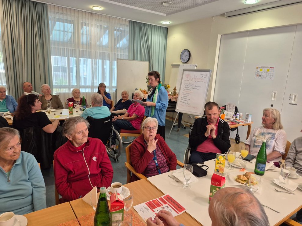 Frau steht und redet. Sitzende Personen beim Tisch.