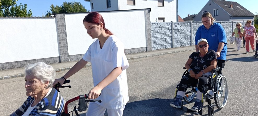Personen gehen spazieren. Praktikantinnen schieben Rollstuhl mit Bewohnerinnen.