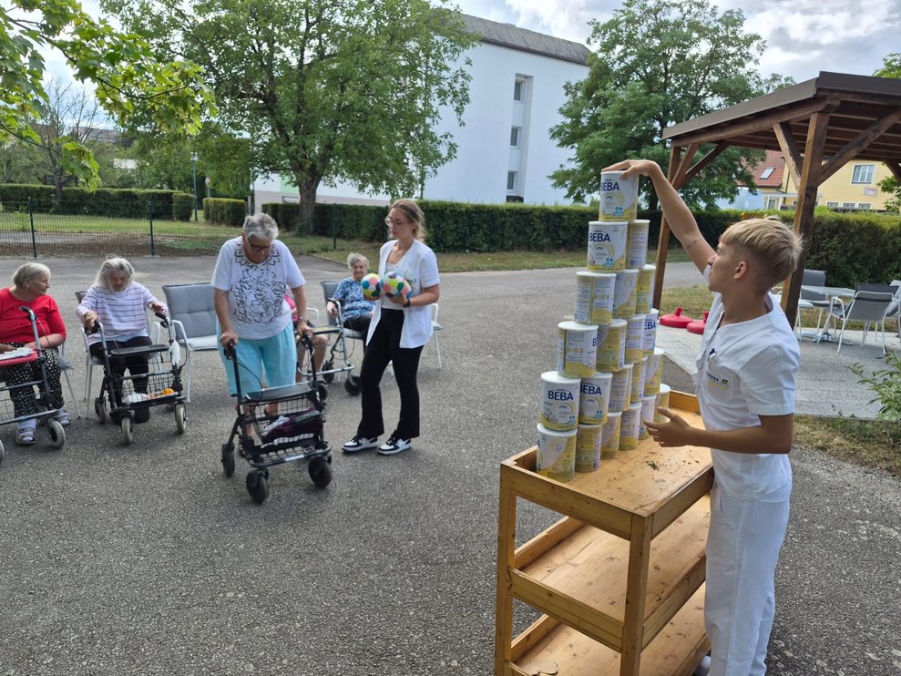 Mann (FSJ) stellt die Dosen auf. Bewohnerin mit Rollator. Praktikantin hält zwei Bälle in den Händen.