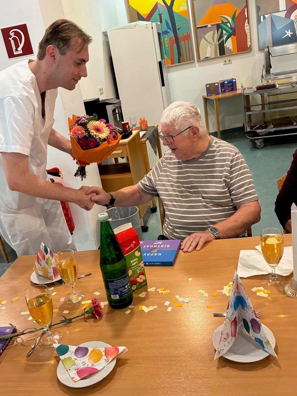 Alltagsbegleiter hält Blumenstrauß in der Hand und gratuliert Bewohner zum Geburtstag. Am Tisch liegt eine Packung Milka Pralinen.