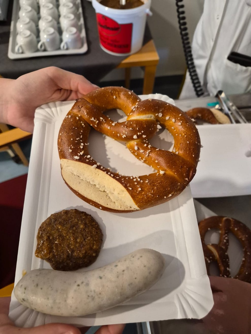 Hand hält kleinen Karton mit Brezen, Weißwurst und Senf.