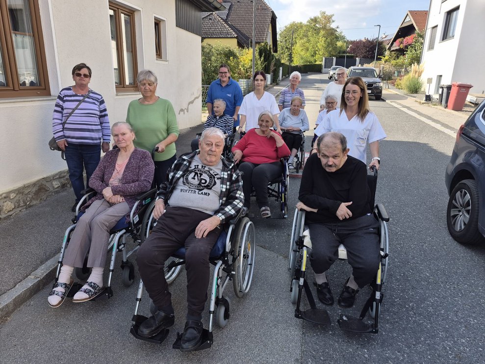 Gruppenfoto beim Spaziergang. BewohnerInnen im Rollstuhl und Personen stehen hinter den BewohnerInnen.