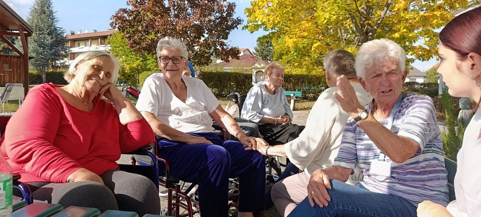 Ehrenamtlich Tätige, Praktikantin und Bewohnerinnen sitzen im Garten und unterhalten sich.