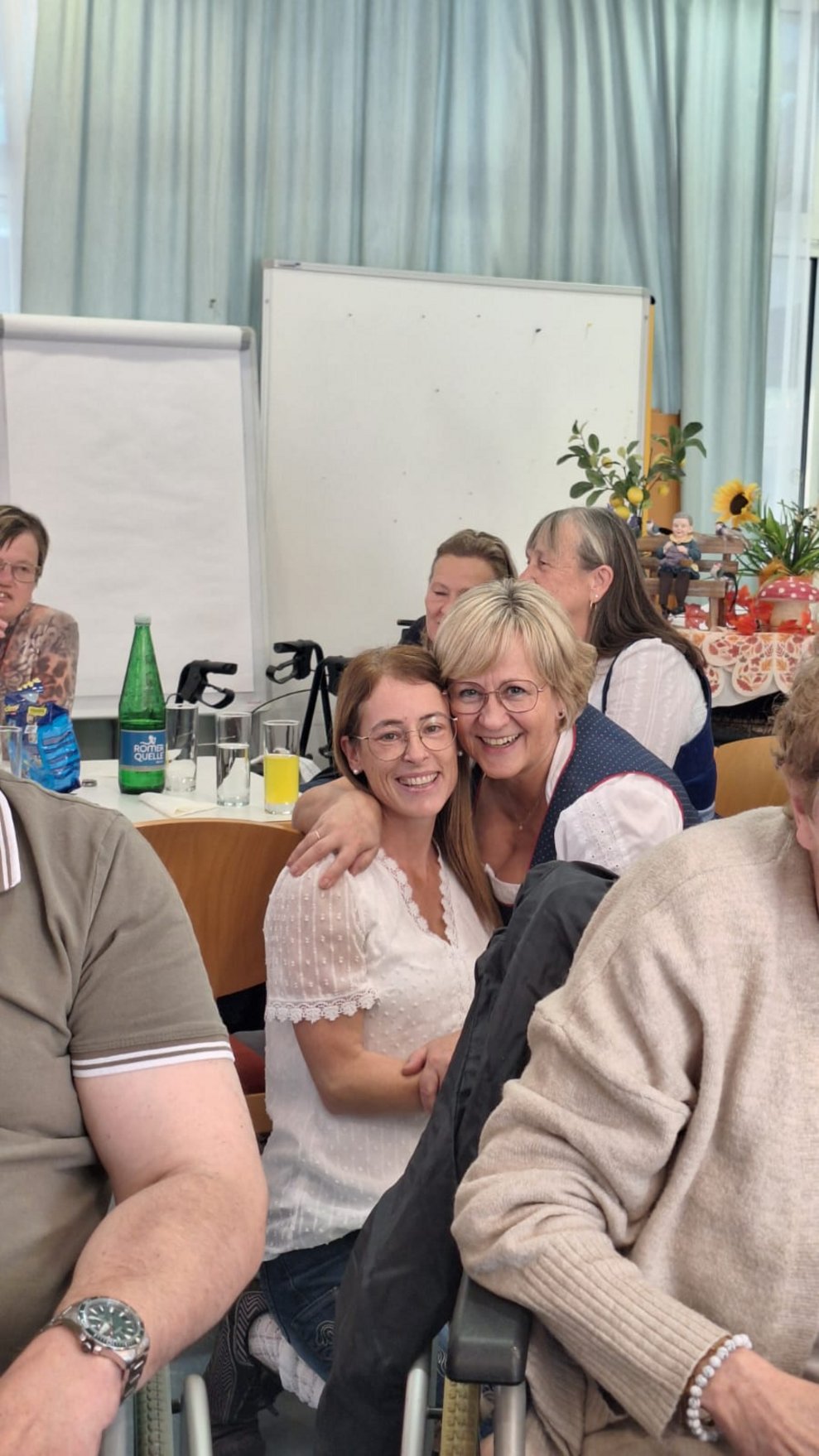 Alltagsbegleiterin und Pflegedirektorin hockerln am Boden und lächeln und halten sich umschlungen.