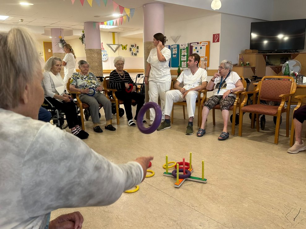 Bewohnerin wirft Ring. Gestell mit Ringen steht auf Boden in der Mitte. Sitzende Personen. Frau (FSJ) steht.