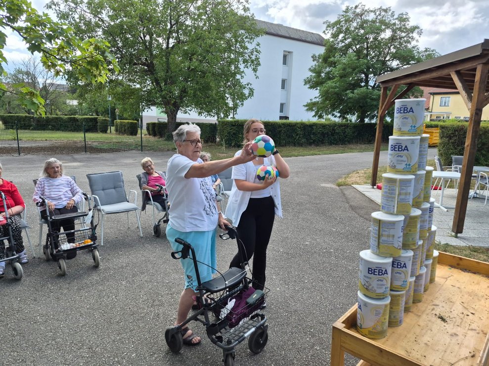 Bewohnerin beim Dosenschießen. Praktikantin steht daneben und hält Ball in der Hand.
