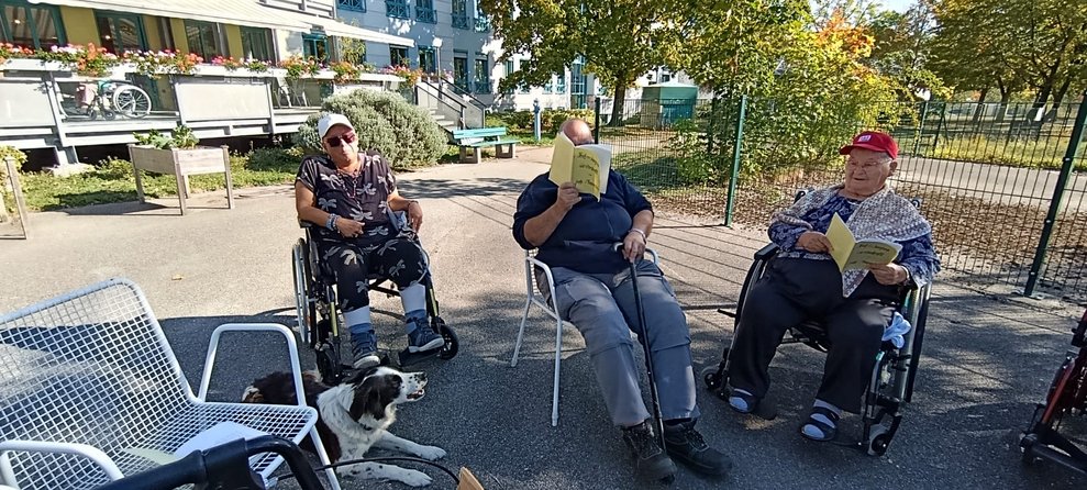 Liegende Hündin. Bewohnerin im Rollstuhl neben Hund. Bewohner und ehrenamtlich Tätiger hält Liederbuch in der Hand.