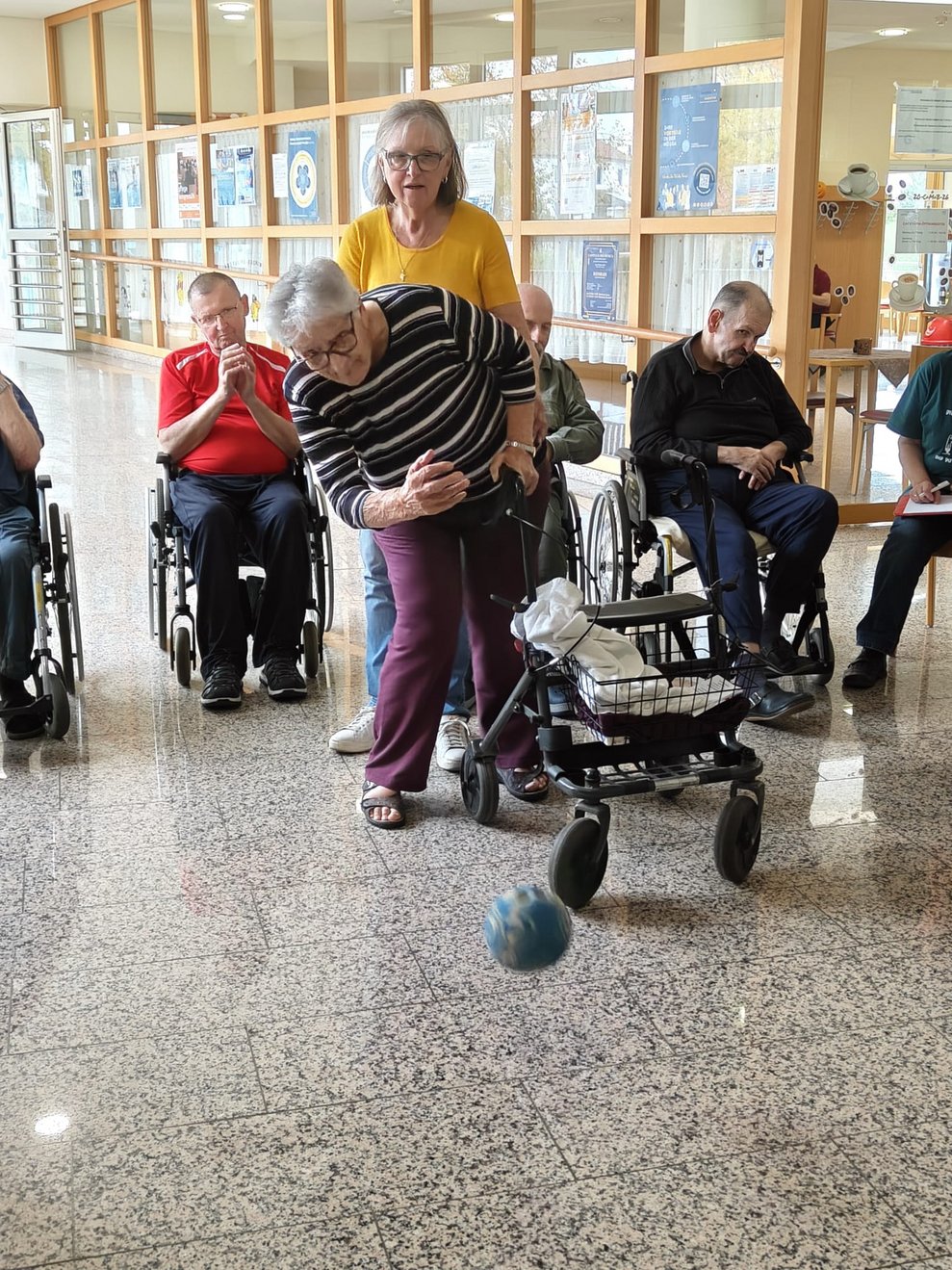 Ehrenamtlich Tätige hält stehende Bewohnerin beim Kegeln. Ball ist in der Luft.