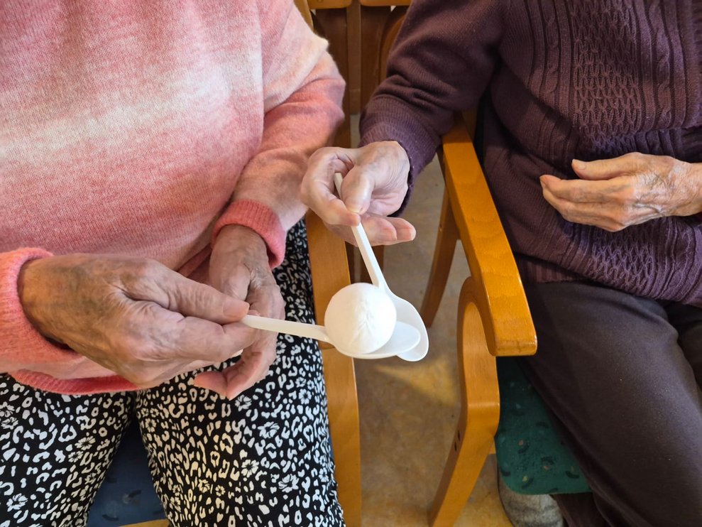 Jeweils zwei Hände halten Kochlöffel und halten kleinen Ball ohne dass er runter fällt. 